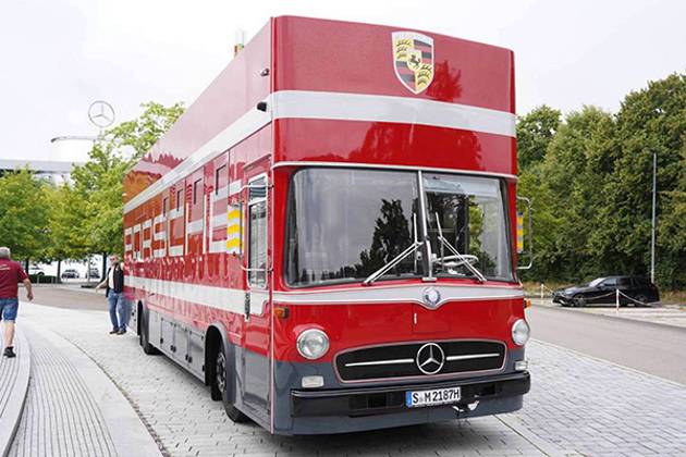 red-mercedes-benz-o317-porsche-logoed-k-race-transporter-on-a-tree-lined-road-mercedes-building-in-the-background