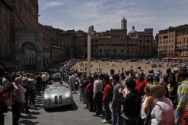 Parade of classic cars driving through a city street with spectators lining the road on a sunny day
