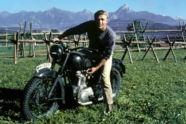 Steve Mcqueen On A Black Triumph Tr6 Motorbike With Wooden Barbed Wire Fencing And A Mountain Backdrop A Scene From The Great Escape