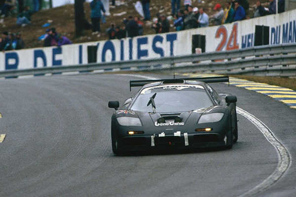 Dark Metallic 1995 Mclaren F1 Gtr Racing Around Racetrack corner with spectators behind an advertising barrier