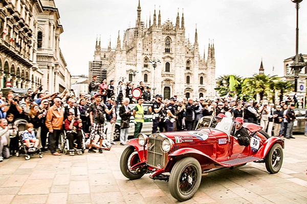 A red classic Alfa Romeo with a crowd and cathedral behind it taking part in the Mille Miglia car race