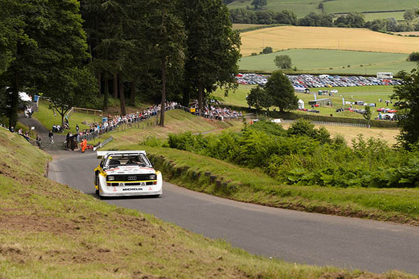 White Audi Sport Quattro S1 Driving Up A Curved Road Back Drop Spectators Lining The Verge Trees Farm Land Fields And Rows Of Parked Cars