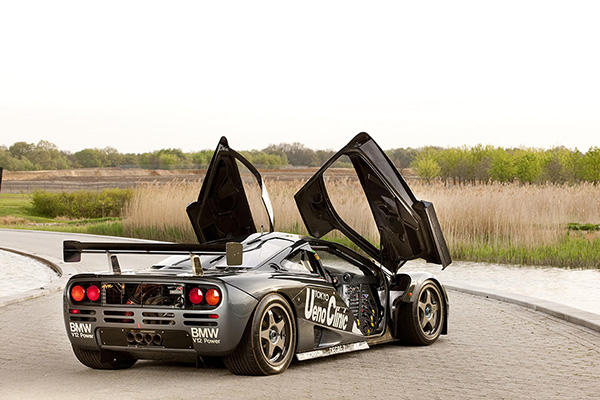 Rear View Of Dark Metallic 1995 McLaren F1 GTR with Doors Up Front View Of Dark Metallic 1995 Mclaren F1 GTR with Doors Up grass and trees backdrop