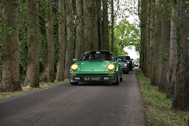 metallic-green-mid-to-late-70s-porsche-911-fronting-a-convoy-of-classic-cars-driving-through-a-single-lane-lined-with-trees