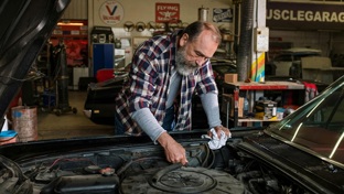 An older man wearing a checked shirt leans over a car engine 