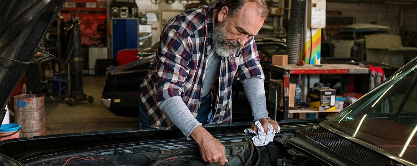 An older man wearing a checked shirt leans over a car engine 