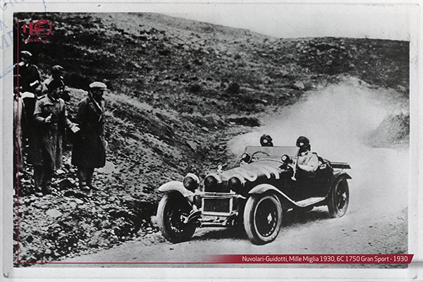 A black and white photo of a claccis car taking part in a race with clouds of dirt behind it on a winding track